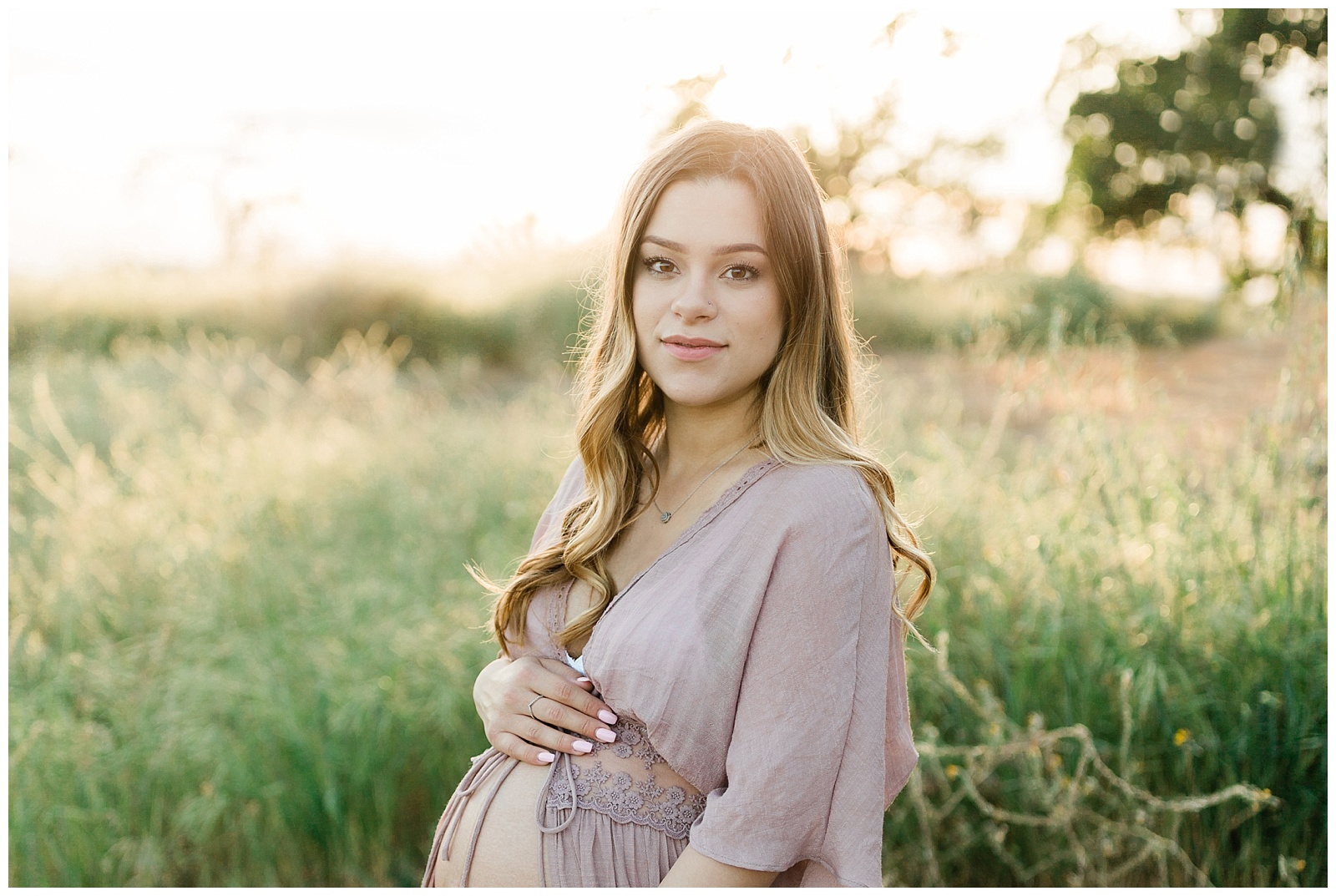  pregnant woman in a grassy field and sunset wearing a blush pink open belly dress