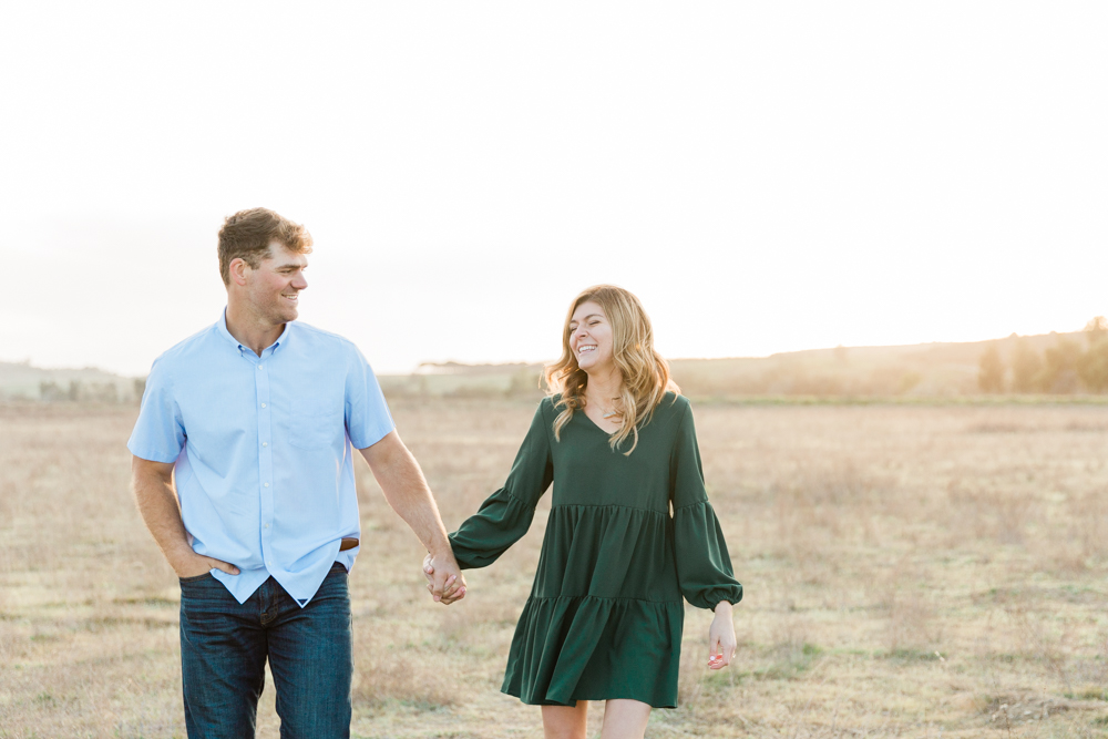 engaged couple walking through an open field at sunset laughing and candid