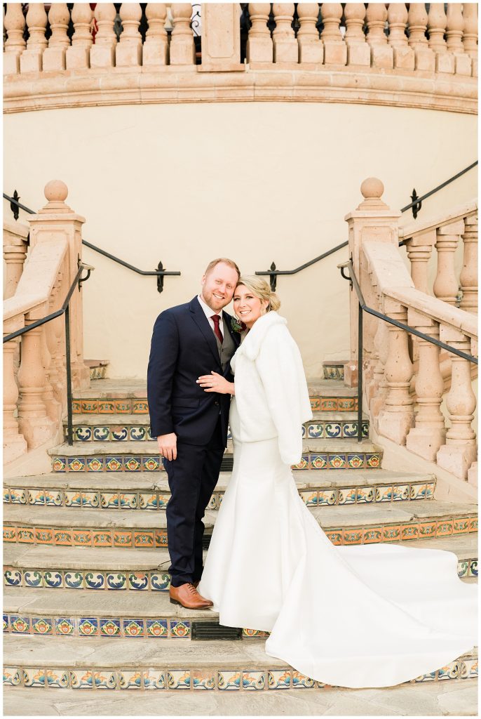 bride in white fur jacket with groom in navy suit on stairs 