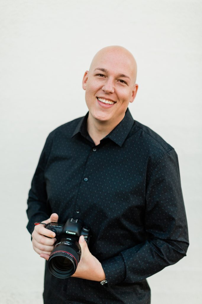photographer standing in front of a white brick wall holding his camera and smiling