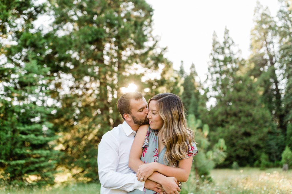 engaged couple hugging in a meadow with her fiance kissing her on the cheek with the sun setting in the backgroun