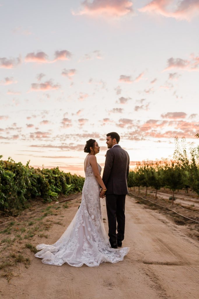 bride and groom holding hands in an orchard under a pink and gold sunset