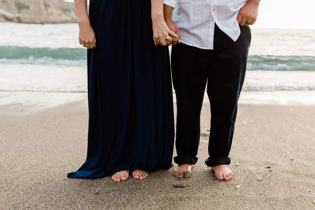 engaged couple holding hands on the beach with their feet in the sand