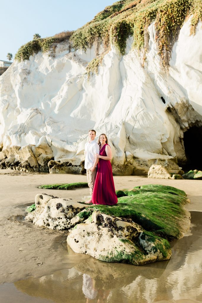 engaged couple standing on a white mossy rock along the shores of pismo beach in california