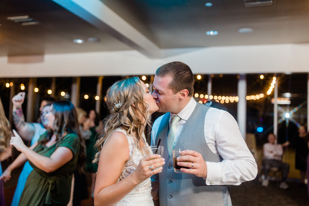 bride and groom holding drinks and kissing at their indoor wedding reception
