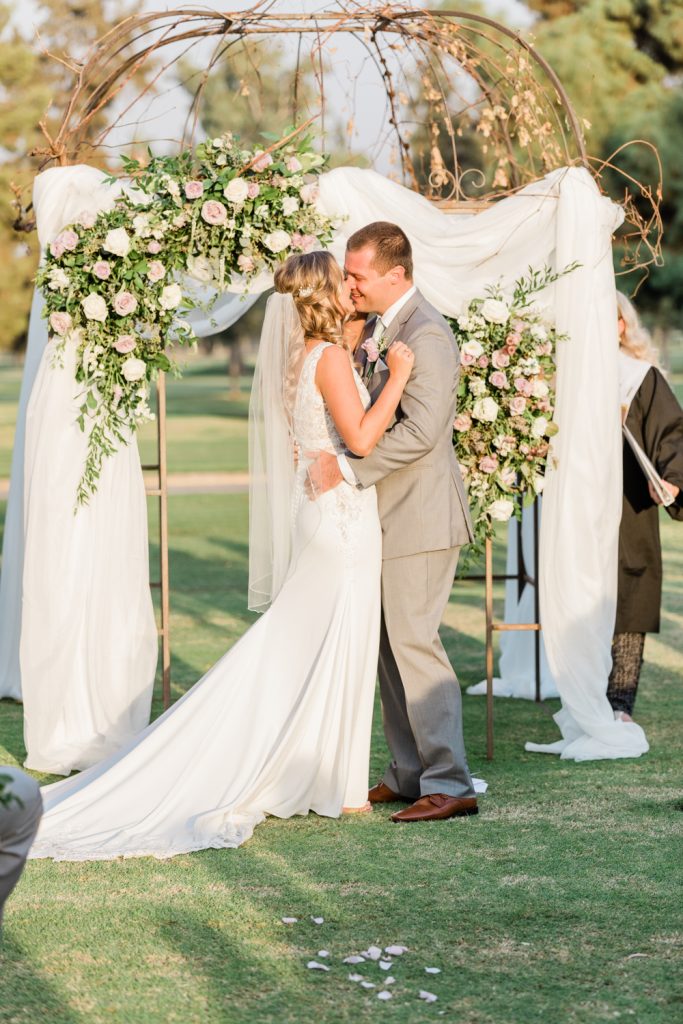 Couples first kiss at outdoor ceremony with draped floral arch.