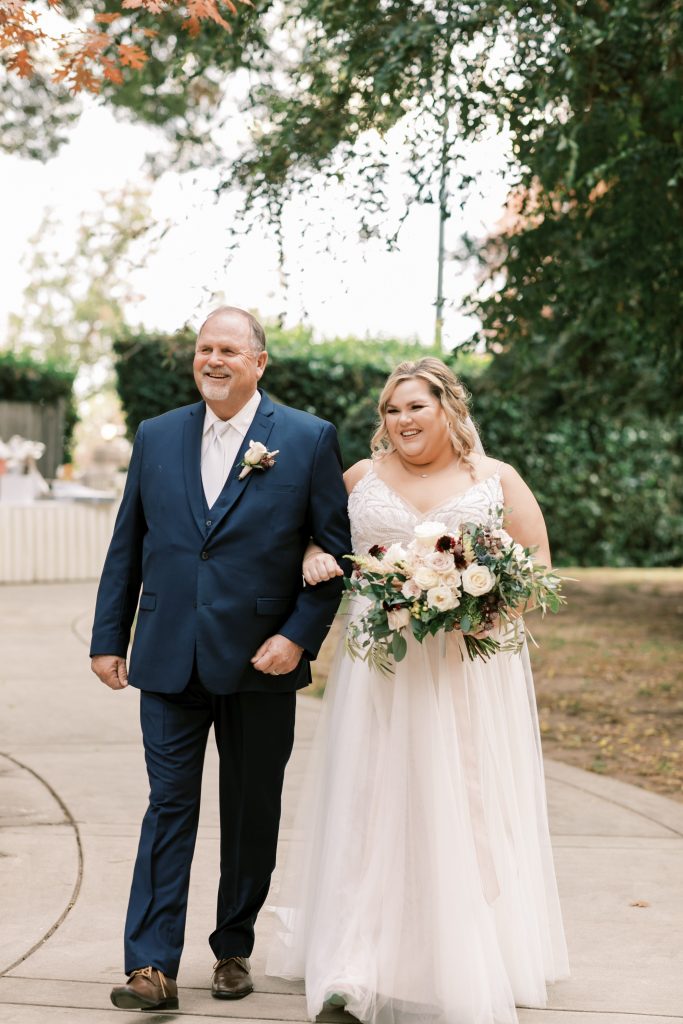 Father walking the bride down the aisle while she smiles on an overcast wedding day