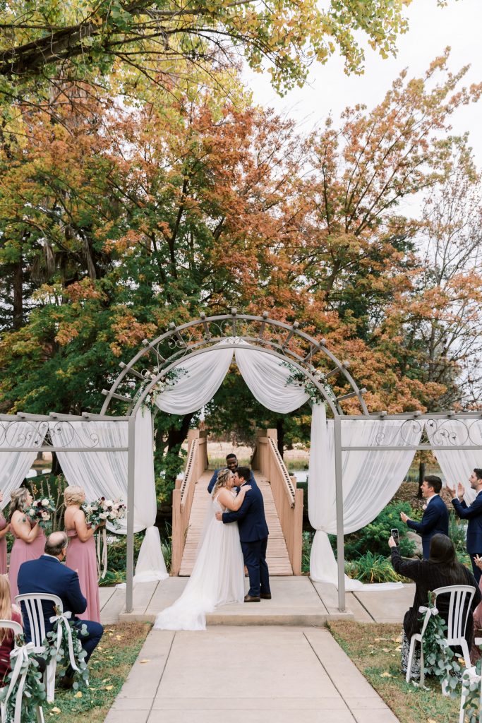 First kiss as husband and wife under draped arch with fall trees behind