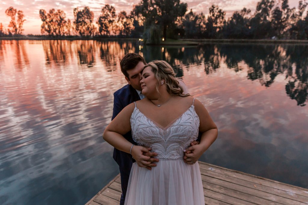 Newlyweds romantically hugging on a dock during a pink sunset