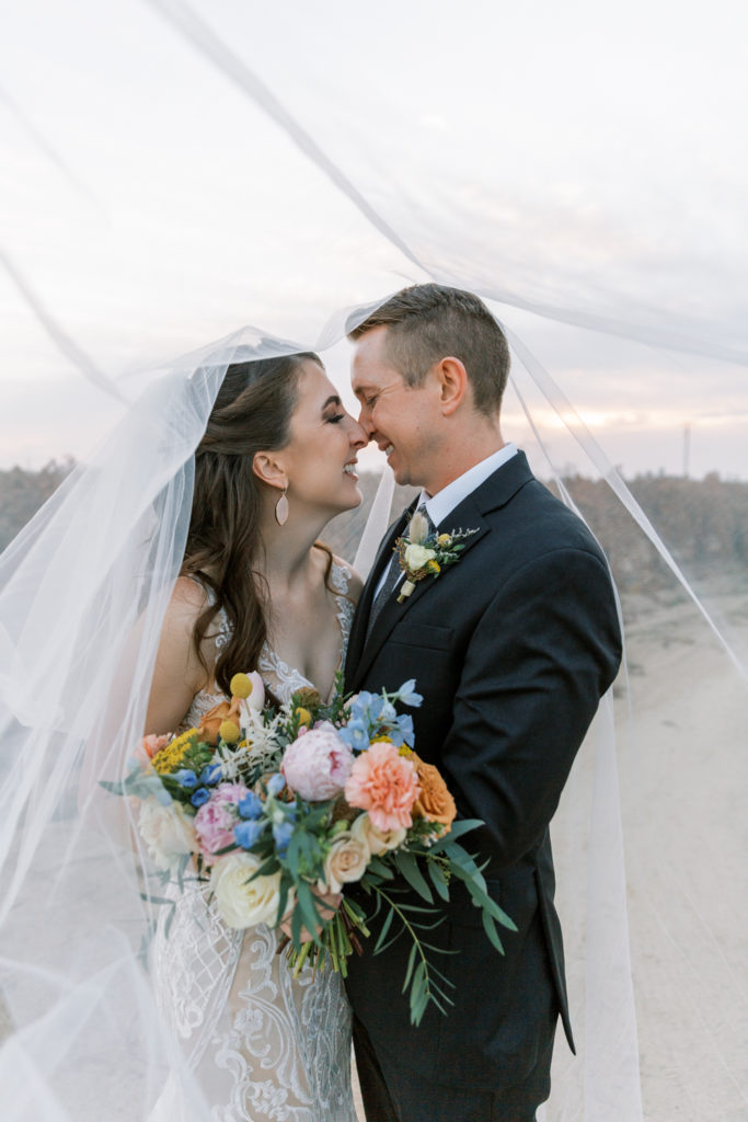 Newlyweds nose to nose under Bride's cathedral veil with colorful bouquet