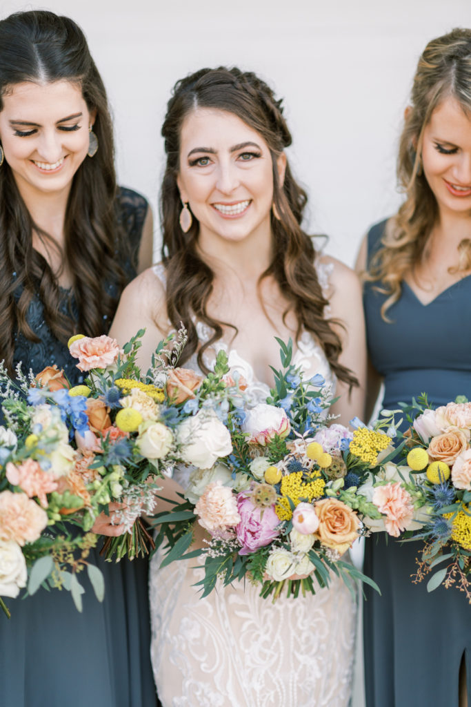 Bride holding colorful bouquet smiling while bridesmaids stand around her. 