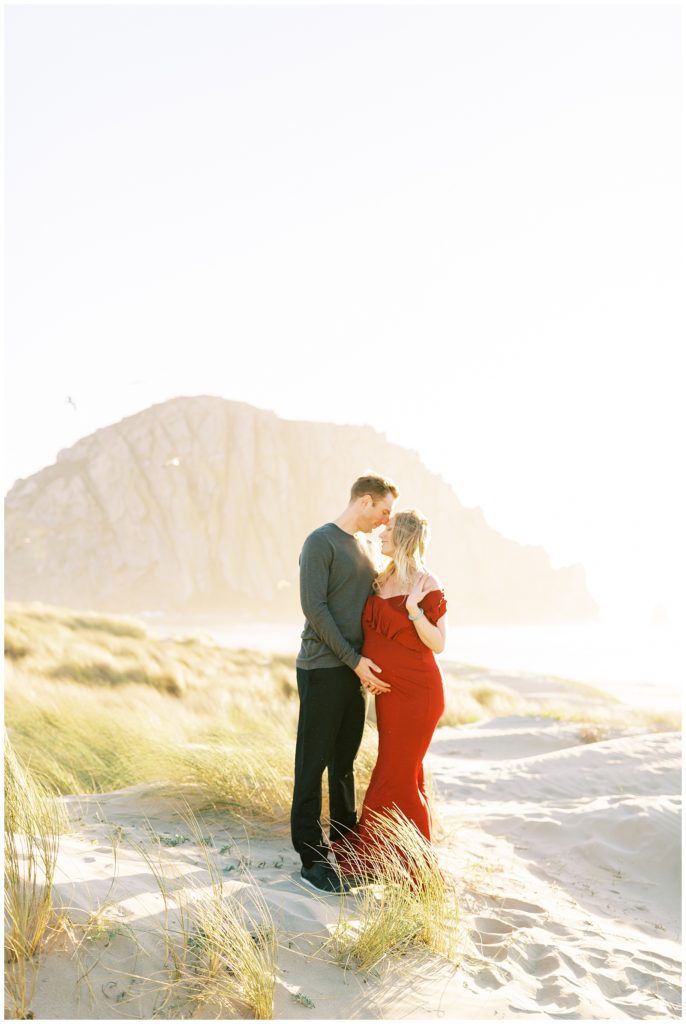 Woman in long red dress with husband for beach maternity photos in morro bay california