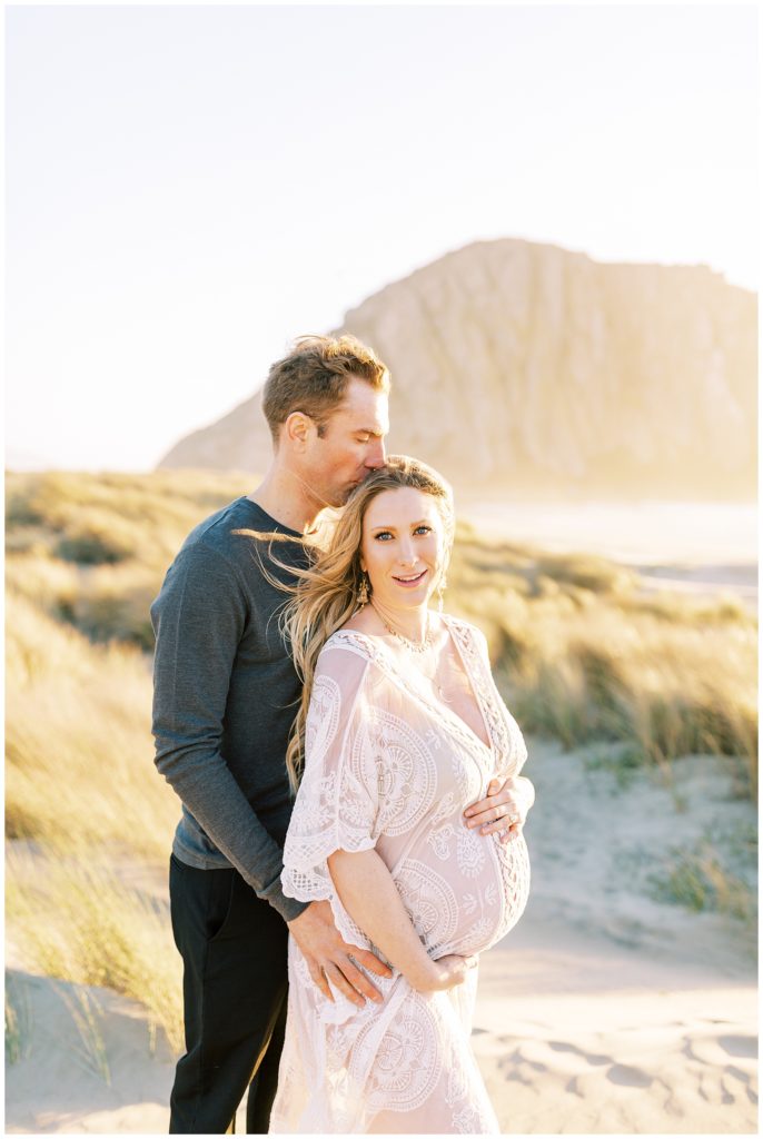Woman holding baby bump and smiling at camera while man kisses her head for beach maternity photos