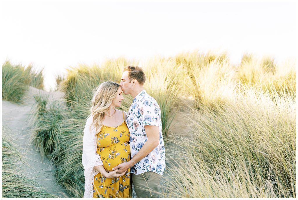 couple in tall grass while man kisses forehead and holds baby bump