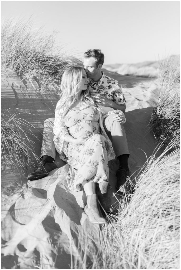 Couple sitting in the sand and grass together lovingly relaxed for beach maternity photos
