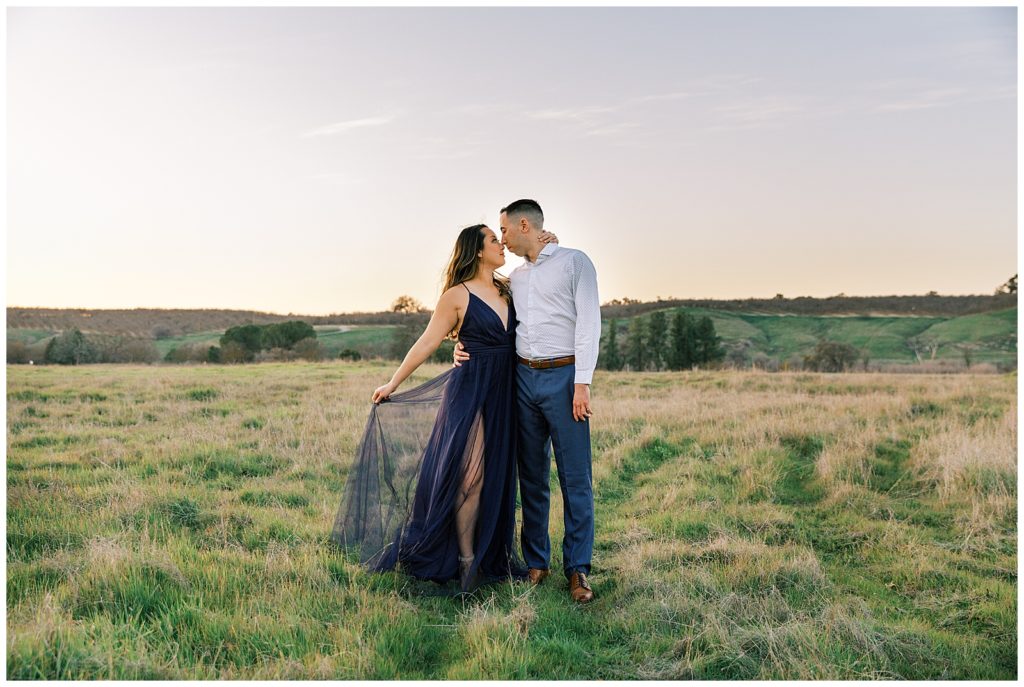 Woman in a chiffon navy blue dress at sunset while fiance hold hers in to him. 