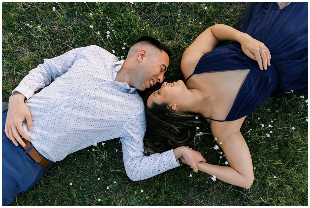 Couple laughing laying on the grass holding hands. 