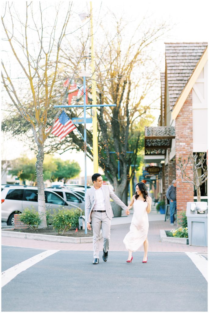 Engaged couple laughing and holding hands while crossing the street in downtown Kingsburg. 