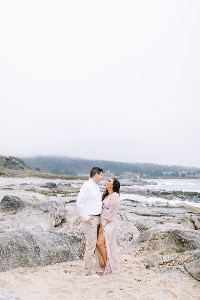 engaged couple looking at eachother and smiling while standing on the rocky beaches of Carmel by the Sea