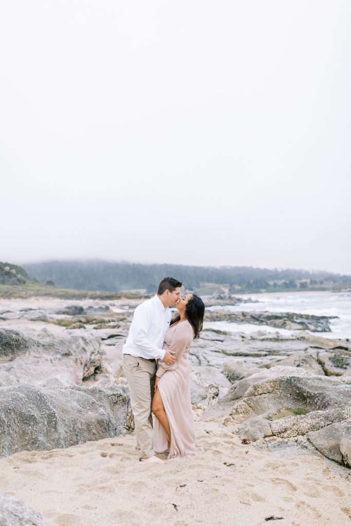 engaged couple standing on a rocky beach in Carmel by the sea california kissing on a foggy day