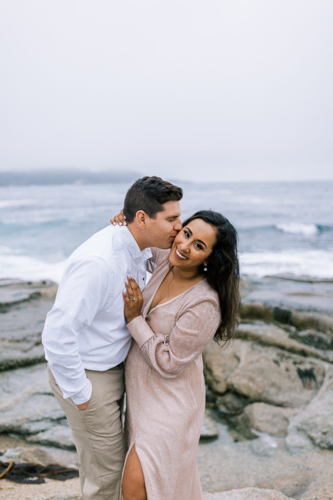 woman smiling while her fiance kisses her on the cheek on a foggy beach in carmel california for carmel engagement photos