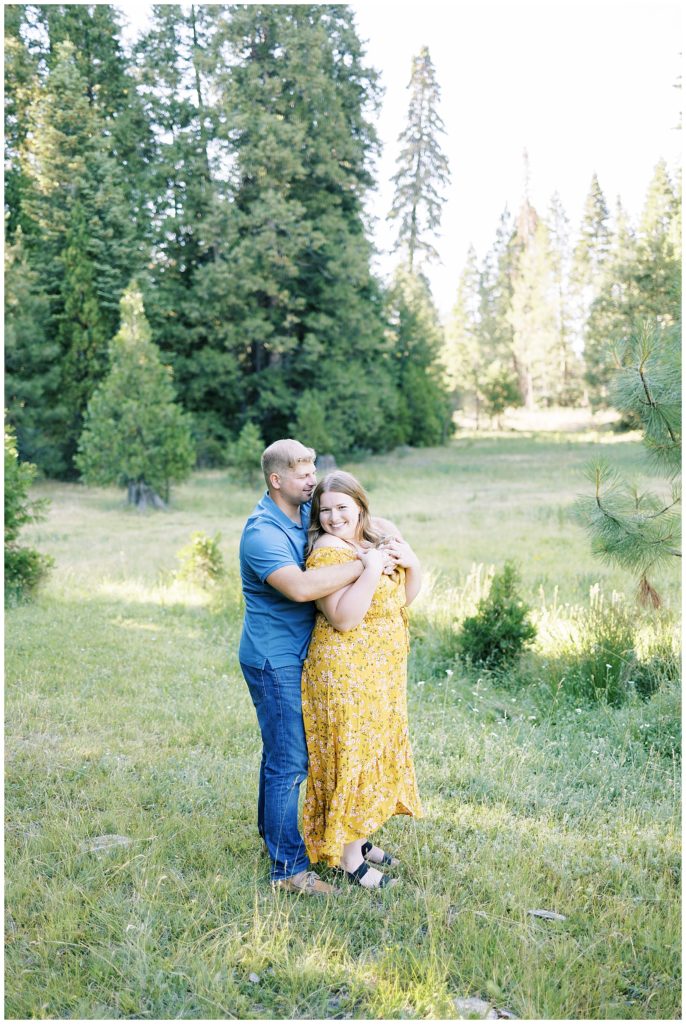 man hugging fiance while woman smiles standing in grassy mountain meadow