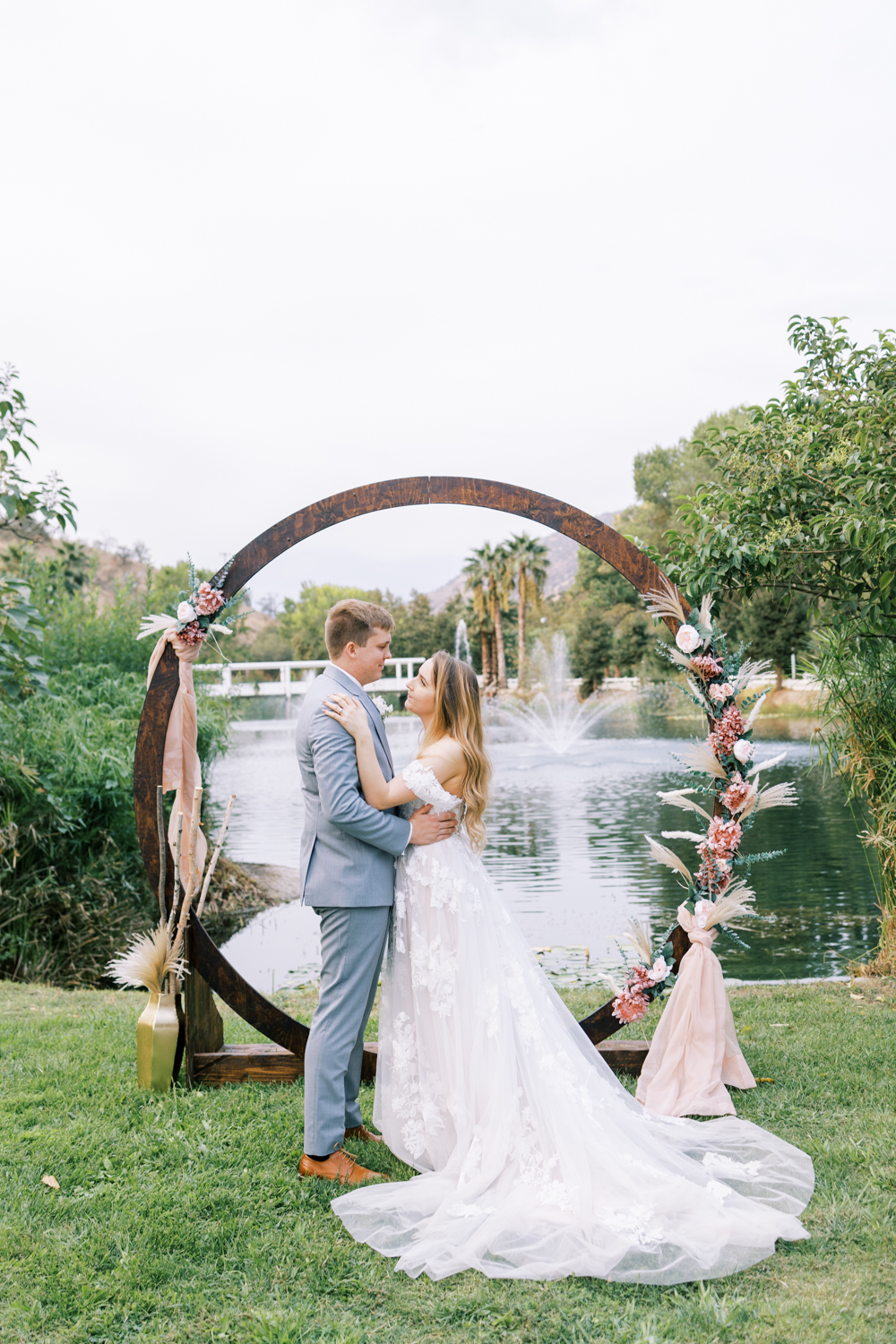 bride and groom standing in front of circle boho wedding ceremony arch in front of a lake