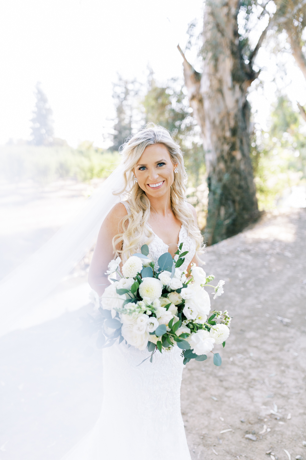 bride holding white and green wedding bouquet and smiling with wedding veil blowing
