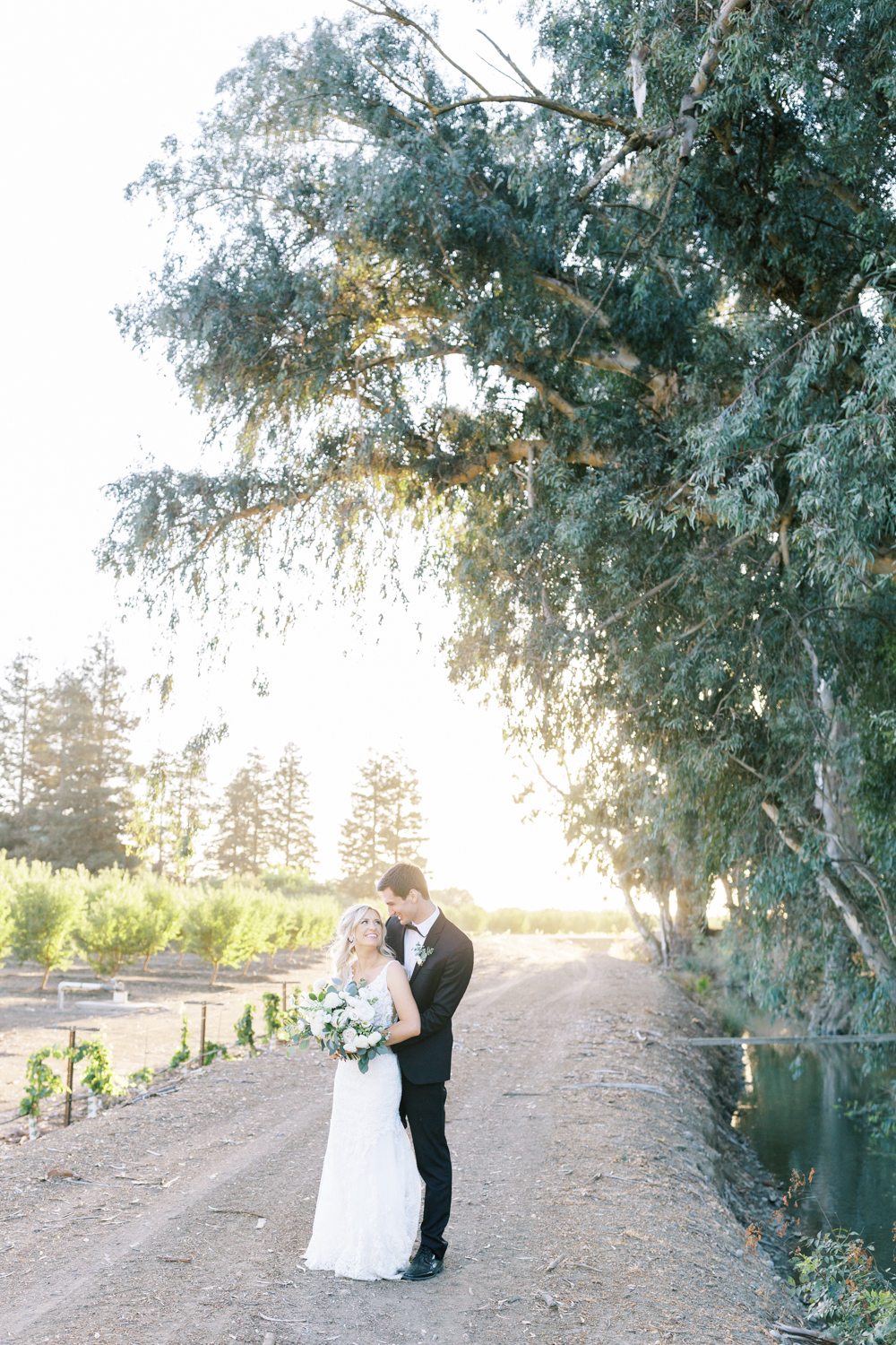groom hugging bride and smiling almond orchard in background along eucalyptus lined canal