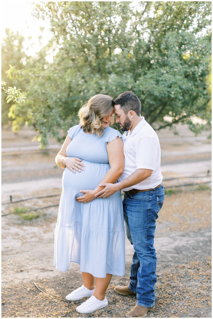 husband and wife embracing while holding baby belly in almond orchard sunlight glowing