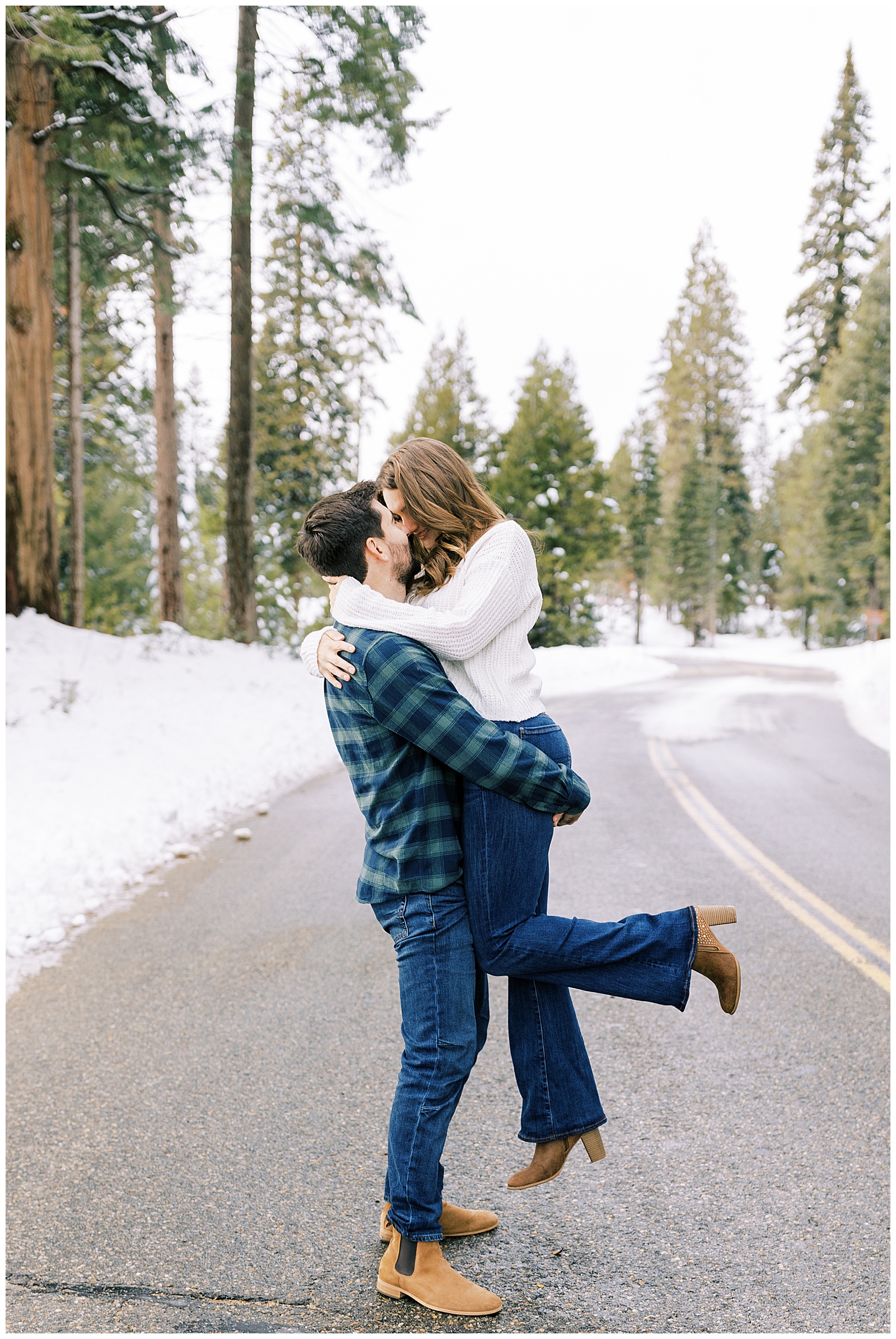 man picking up fiance kissing her on mountain road evergreen trees snow winter engagement photos