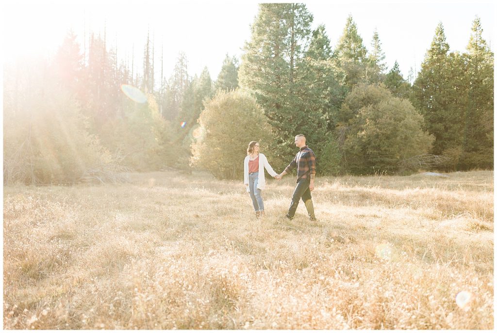 engaged couple holding hands and walking through a dried grassy meadow in the mountains of California