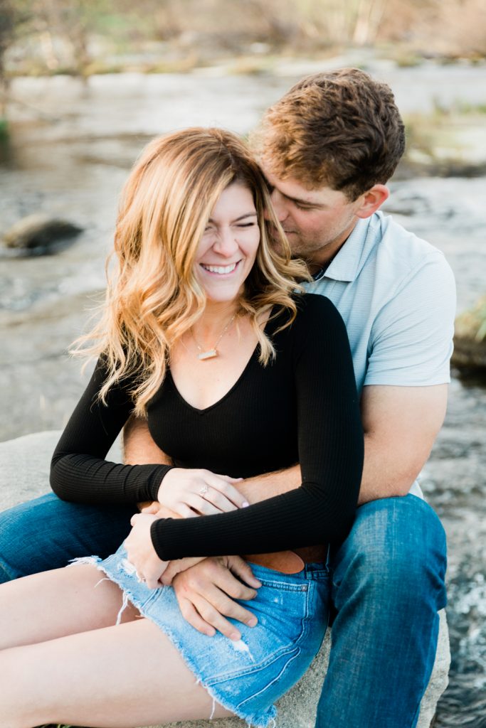 candid moment of a newly engaged couple cuddling on a rock on the edge of a river