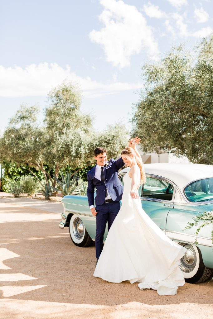 groom in navy suit twirling bride in front of vintage car