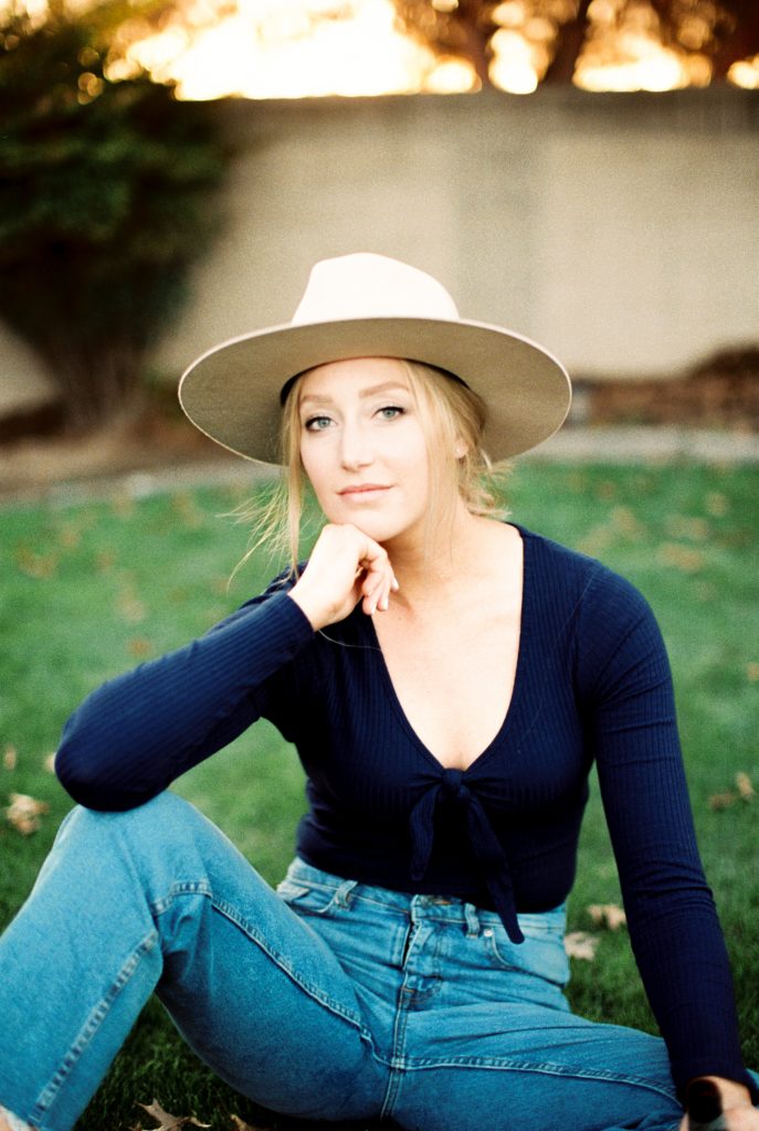 blonde woman wearing a beige suede hat and a navy blue shirt sitting in the grass