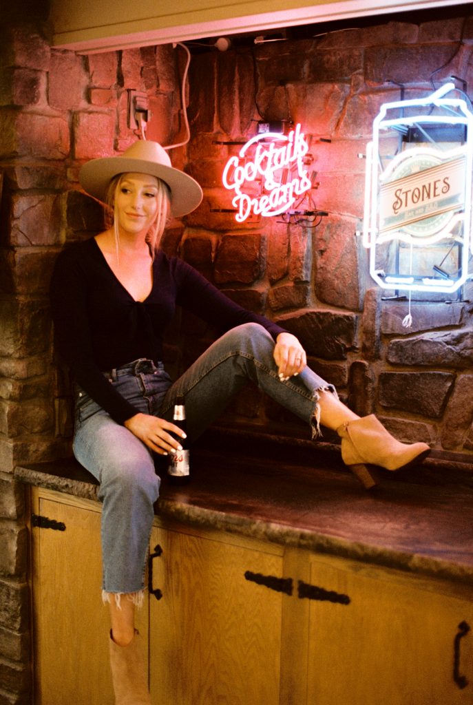 woman sitting on a bar with neon signs holding a coors light