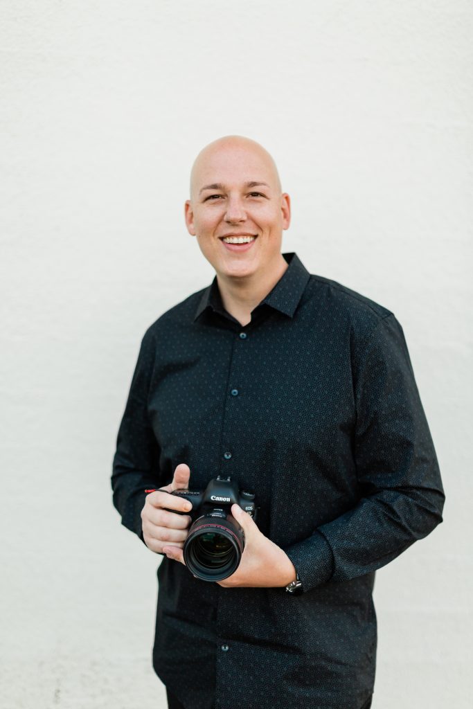 man smiling and holding a canon camera while wearing a black button down shirt