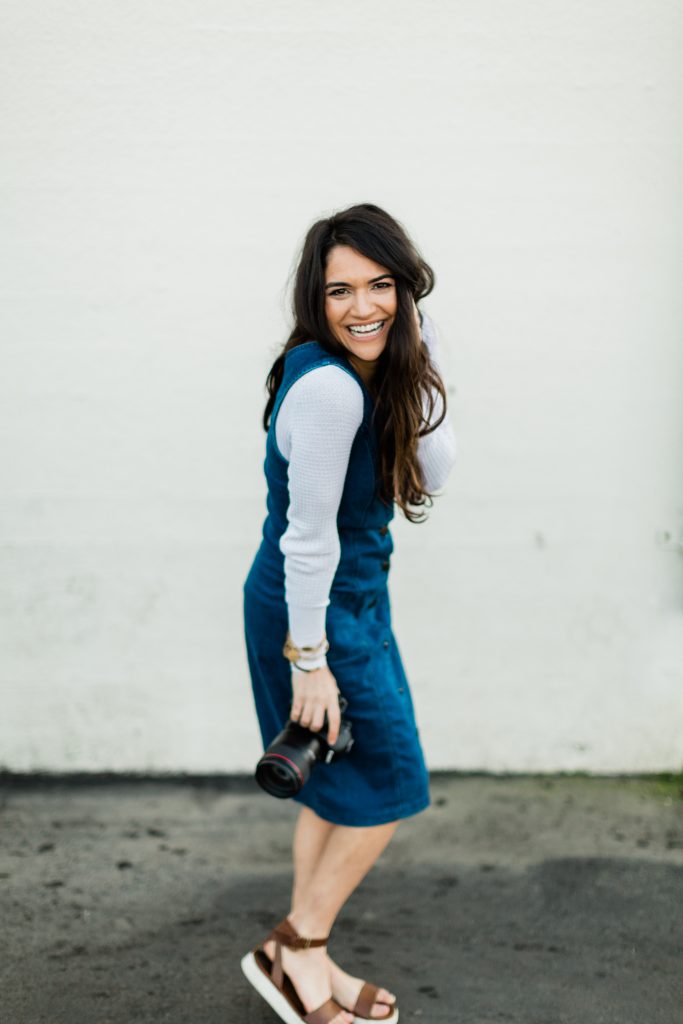 photographer smiling in front of a white brick wall in a denim dress holding her camera