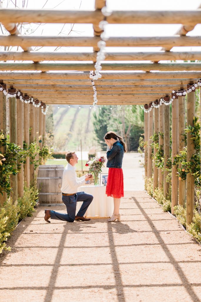 woman covering her face in surprise as her boyfriend surprises her with a proposal under a wooden archway
