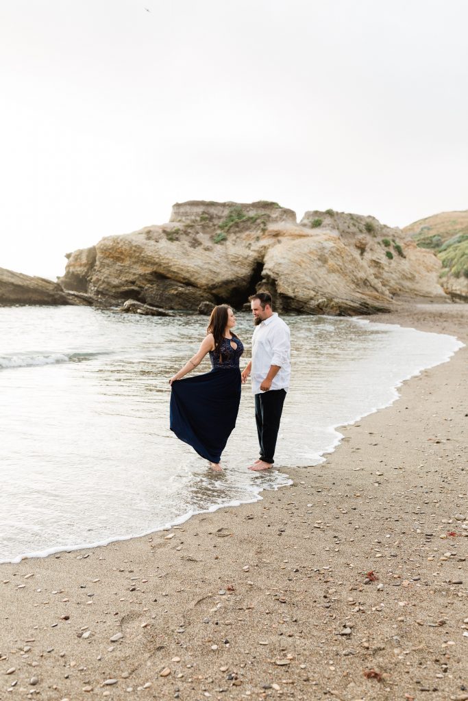 engaged couple standing along the shores of montana de oro beach in a navy blue maxi dress