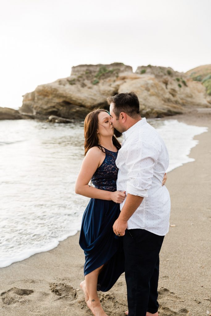 engaged couple kissing on the shores of montana de oro and the waves come up and touch their feet