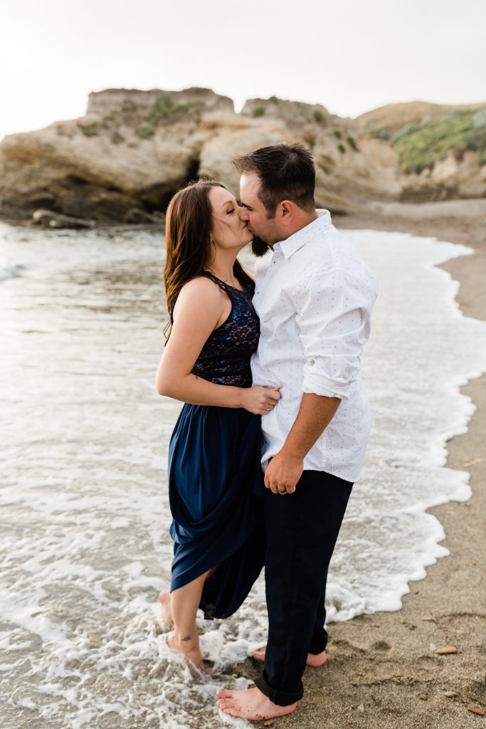 engaged couple kissing in the waves on the shores of montana de oro wearing a navy blue dress for the girl and black pants and a white button down shirt for the guy