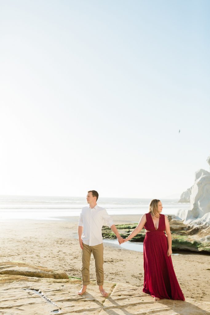 couple holding hands and standing on rocks along the beach of pismo beach