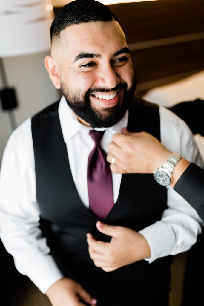 groom smiling while someone fixes his maroon tie