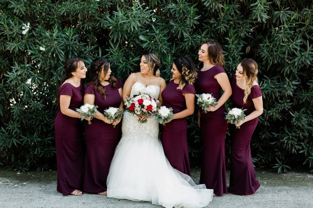bride and bridesmaids in maroon maxi dresses holding white and eucalyptus bouquets while standing in front of an oleander bush at r wedding house