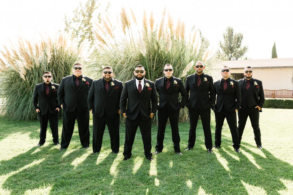 groom and groomsmen standing in a v formation wearing black calvin klein suits from mens wearhouse with sunglasses on and pampas grass in the background