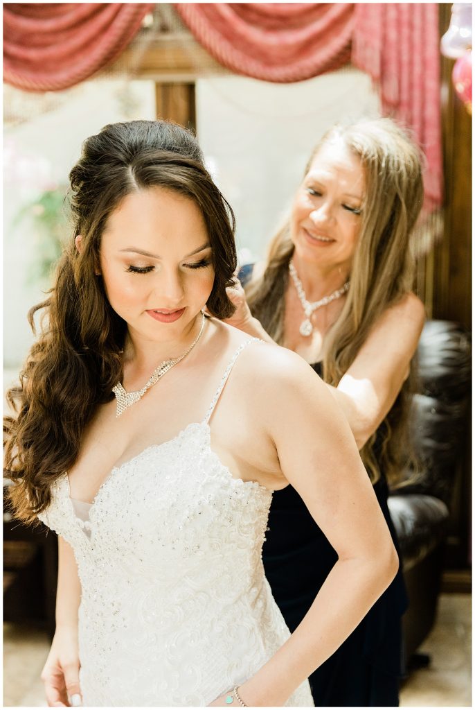 Bride in beaded spaghetti strap dress having her mom put her necklace on