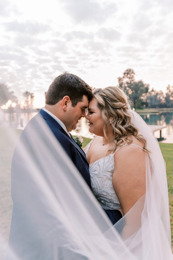 Bride and groom nuzzling during sunset with veil flowing in front.