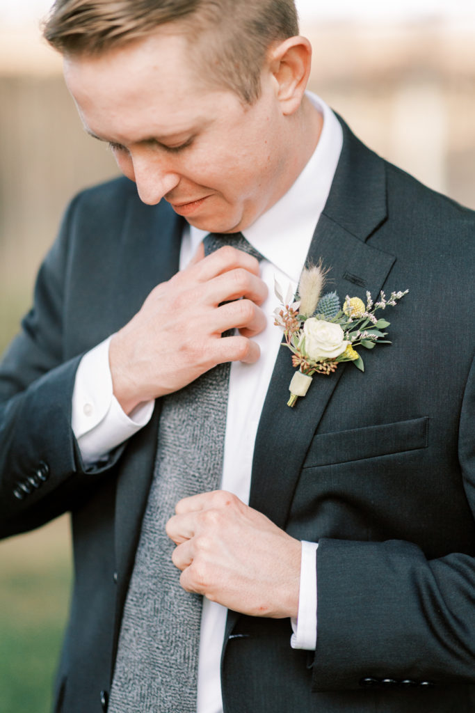 Groom fixing grey tie while wearing a charcoal suit and with a white rose boutonniere 