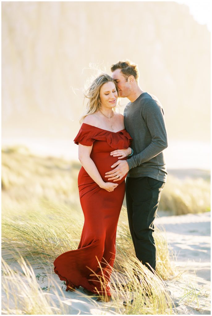 Husband whispering to pregnant wife while they stand in the grass in Morro Bay by california photographer megan helm
